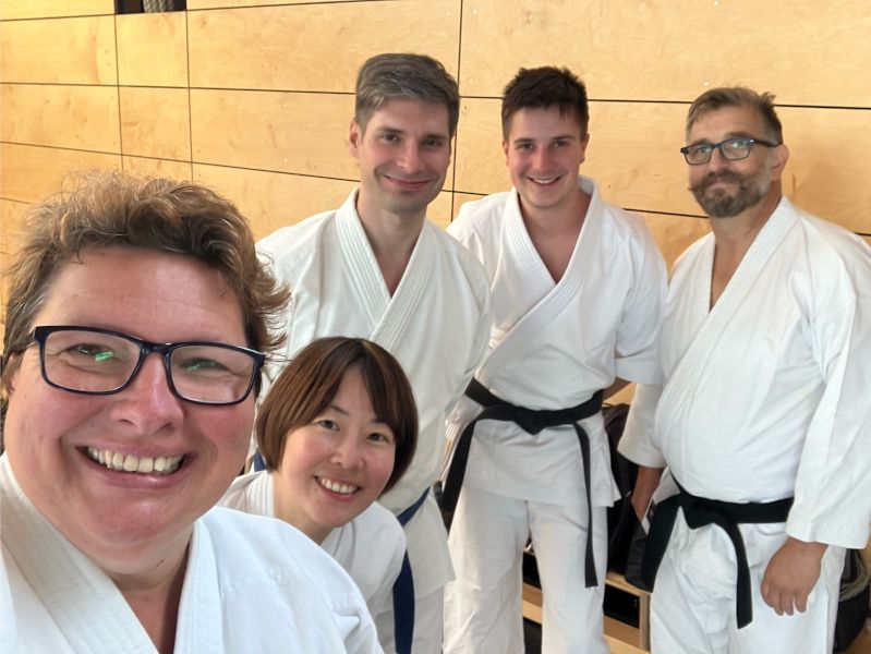 Fünf Karateka stehen vor einer holzvertäfelten Wand in einer Turnhalle und lächeln in die Kamera. Alle fünf haben Karate-Gis an.