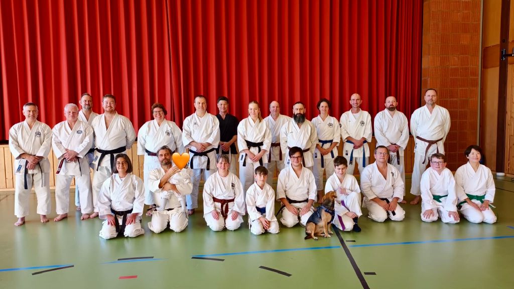 Etwa zwanzig Karateka stehen und sitzen auf dem Boden einer Turnhalle vor einem Vorhang. Ein Hund sitzt vor der Gruppe auf dem Boden.