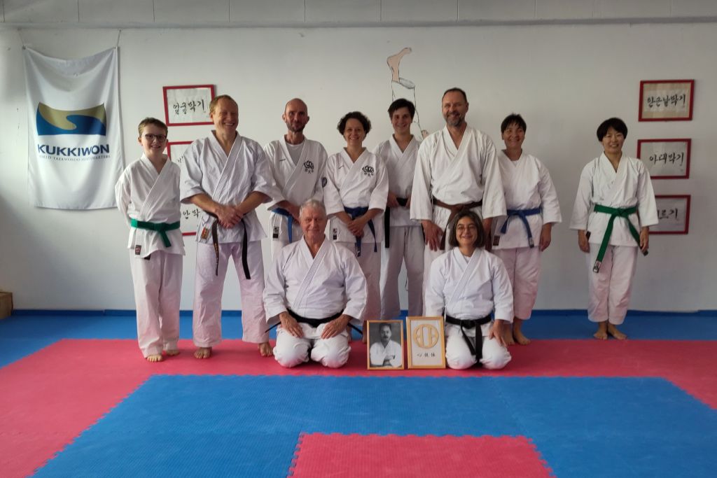 Acht Karateka stehen in einem mit Gummimatten ausgelegten Raum vor einer Wand. Vor ihnen sitzen zwei weitere Karateka im Seiza. Zwischen den beiden sind ein Schwarz-Weiß-Foto eines Mannes und das Familienwappen der Familie Mabuni jeweils in einem Bilderrahmen aufgestellt.