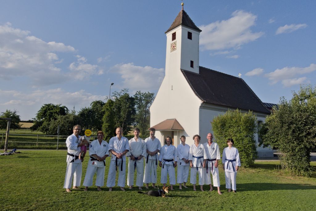 Zehn Karateka stehen auf einer Wiese und schauen zur Kamera. Hinter ihnen steht eine Kapelle. Vor ihnen auf der Wiese liegt ein Hund.