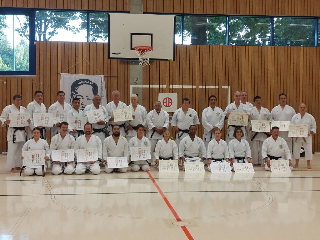 Fünfundzwanzig Karateka sind in zwei Reihen in einer Turnhalle aufgereiht. Die vordere Reihe kniet im seiza, die hintere Reihe steht. Ein Großteil hält eine große Urkunde mit japanischen Schriftzeichen hoch.