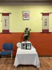 Das Shōmen in der Haupt-Trainingshalle. Auf einem Tisch stehen zwei eingerahmte Fotos, die Mabuni Kenei Sōke (links) und Mabuni Kenwa Sōke (rechts) zeigen. Hinter den Fotos steht eine Vase mit Blumen. An der Wand hängen zwei Schriftrollen mit japanischen Zeichen, links „空手道“ (karate-dō), rechts „心技体“ (shin gi tai). Dazwischen hängt ein Bild mit dem Familiensymbol der Familie Mabuni, ein Kreis mit zwei vertikalen Linien, die nach rechts und links mit kurzen horizontalen Linien zum Kreis verbunden sind, und den Schriftzeichen „糸東流“ (shitōryū).