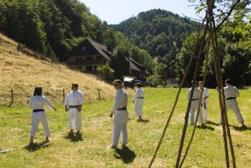 Auf einer Wiese unterhalb eines Hangs trainieren Karateka paarweise. Die Sonne scheint.