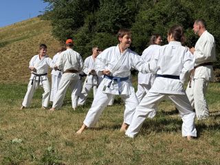 Aufwärmen, Trainingseinheit Manuel Auf einer Wiese unterhalb eines Hangs stehen Karateka paarweise beieinander. Sie haben jeweils am Unterarm Kontakt zueinander und bewegen ihre Schwerpunkte synchron und abwechselnd vor und zurück.