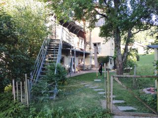 Blick über ein Gartentor auf die Rückseite eines mehrstöckigen Hauses. Vor dem Haus stehen Bänke und Tische.