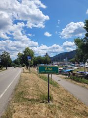 Ein grünes Ortsschild mit gelber Schrift steht zwischen dem Straßenrand und einem Rad- und Fußweg. Auf dem Schild steht „Aha“ und darunter „Gde. [Gemeinde] Schluchsee.“ Im Hintergrund stehen auf einer Wiese kleine Segelboote, weiter Weg ist ein See zu sehen. Die Sonne scheint.